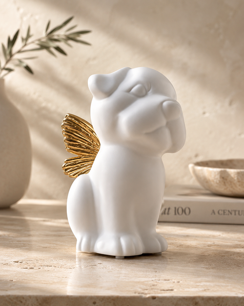 A white ceramic angel dog figurine with a metallic gold wing, displayed on a beige stone surface with a neutral background and soft shadows.