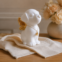 A white ceramic angel dog figurine with closed eyes, small gold wings, and a gold necklace with a round pendant, displayed on a beige cloth on a wooden table with books and flowers in the background.